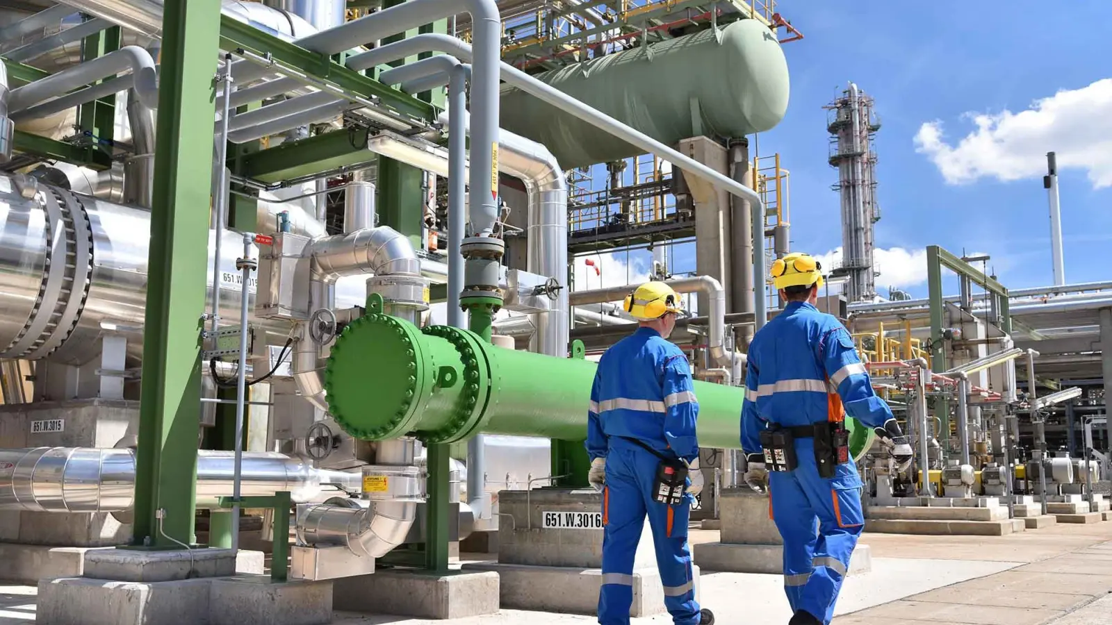 Operarios caminando por una planta de procesos con tuberías industriales y equipos de intercambio de calor