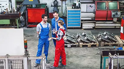 Técnicos en un taller industrial conversando junto a palés con piezas metálicas